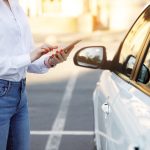 Close up woman using mobile phone, communication or online application, standing near car on city street or parking, outdoors. Car sharing, rental service or taxi app.