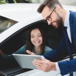 Male handsome caucasian shop assistant showing explaining to a female client customer car options information on digital tablet before buying choosing new car auto